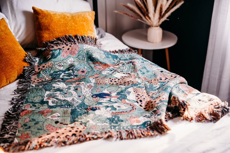 May include: A green and brown woven throw blanket with a fringed edge features a pattern of leopards and jungle foliage. The blanket is draped over a white bed with orange pillows.