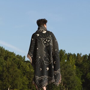 May include: A person wearing a black woven tapestry blanket with a cat and moon design. The blanket has fringe on the edges. The person is walking in a grassy field with trees in the background.