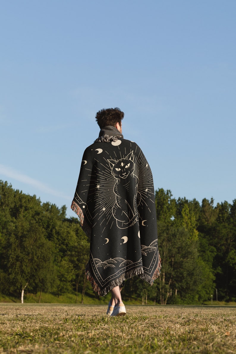 May include: A person wearing a black woven tapestry blanket with a cat and moon design. The blanket has fringe on the edges. The person is walking in a grassy field with trees in the background.