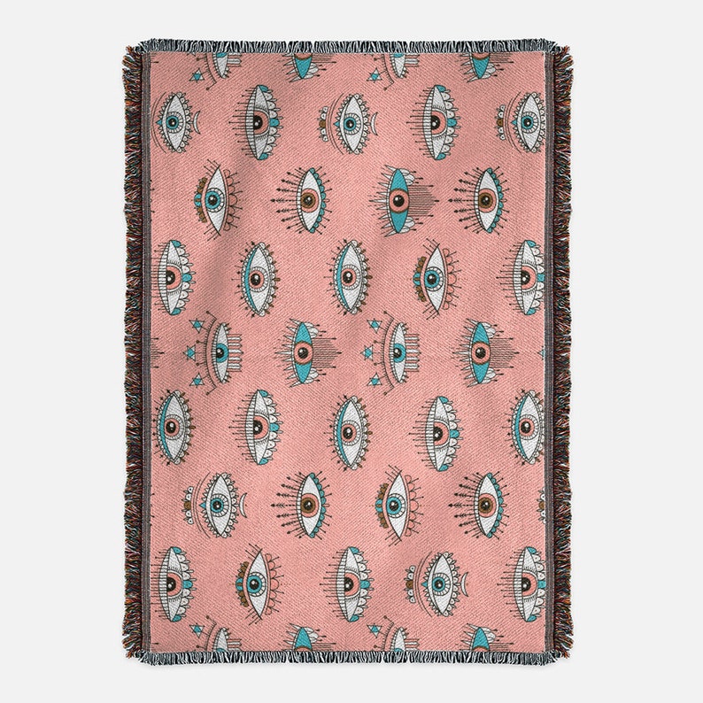 K&ouml;nnte beinhalten: Eine rosa gewebte Wolldecke mit einem sich wiederholenden Muster aus blauen und wei&szlig;en stilisierten Augen. Die Augen haben ein dekoratives Design um sie herum.