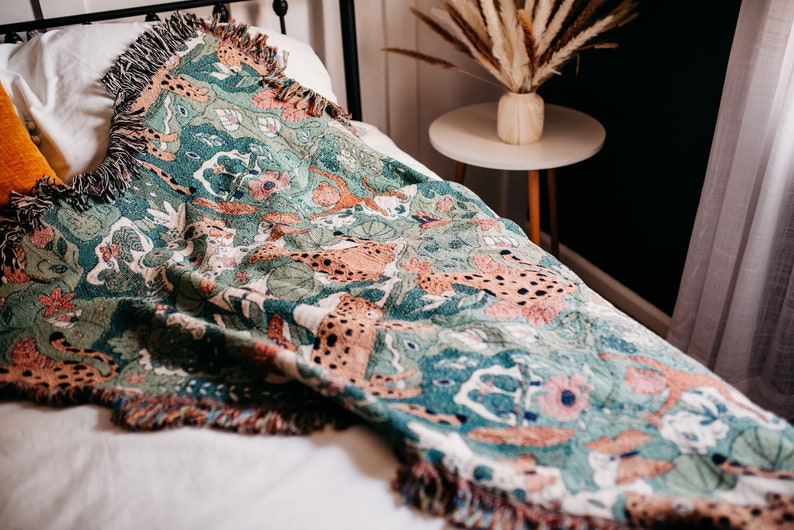 May include: A green, orange, and white woven throw blanket with a pattern of leopards and flowers. The blanket is draped over a bed with a white pillow and a yellow pillow.