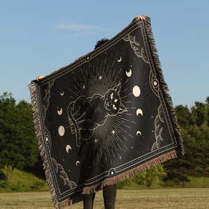 May include: A black woven tapestry with a gold design of a cat with three eyes, surrounded by moons and stars. The tapestry is being held up by a person in a grassy field.