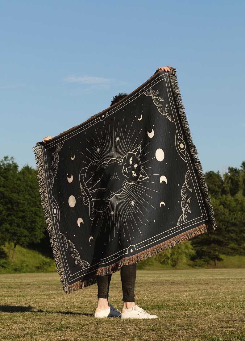 May include: A black woven tapestry with a gold design of a cat with three eyes, surrounded by moons and stars. The tapestry is being held up by a person in a grassy field.