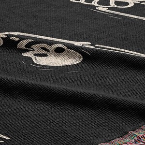 May include: Black woven throw blanket with a white skeleton design. The blanket has a fringe border.