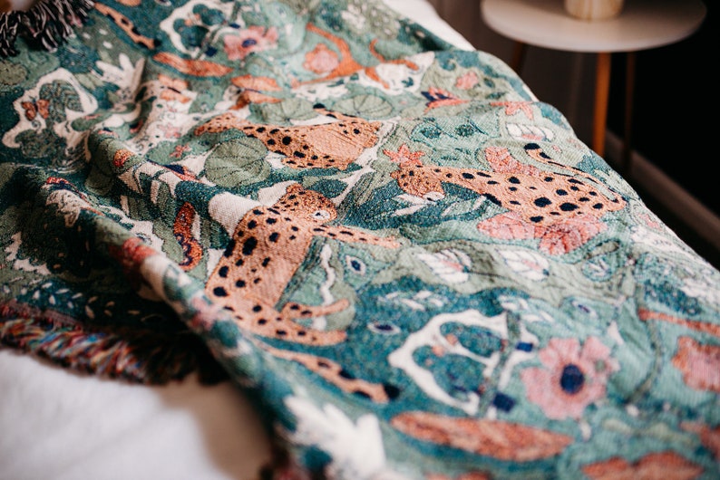 May include: A woven throw blanket with a green background and a pattern of orange and brown leopards. The blanket has a fringed edge.