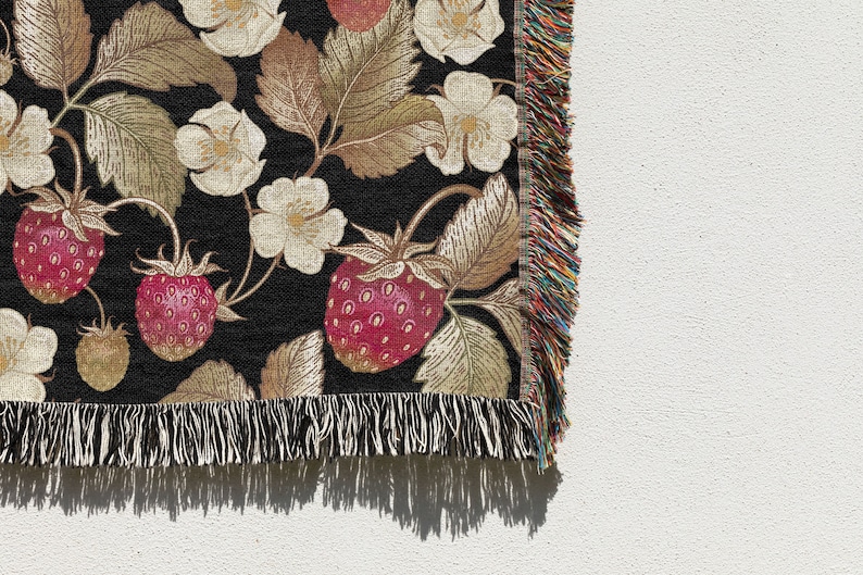 May include: A woven throw blanket with a black background featuring a strawberry and floral pattern. The strawberries are red, and the flowers are white. The blanket has a fringed edge with multiple colors.