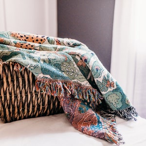 May include: A woven basket filled with a colorful throw blanket with a green, orange, and brown pattern. The blanket has fringe on the edges.