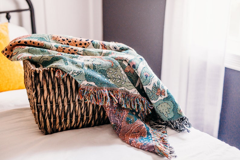 May include: A woven basket filled with a colorful throw blanket with a green, orange, and brown pattern. The blanket has fringe on the edges.