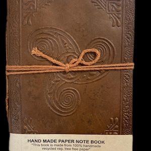 May include: A brown leather-bound journal with a Celtic knot design on the front cover. The journal is tied closed with a brown twine. The label on the front reads "HAND MADE PAPER NOTE BOOK" and "This book is made from 100% handmade recycled rag, tree free paper".