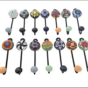 May include: A collection of fourteen wall hooks featuring black metal stems and round ceramic knobs. Each knob displays a unique hand-painted pattern in various colours including blue, red, yellow, green, orange, black, and white.