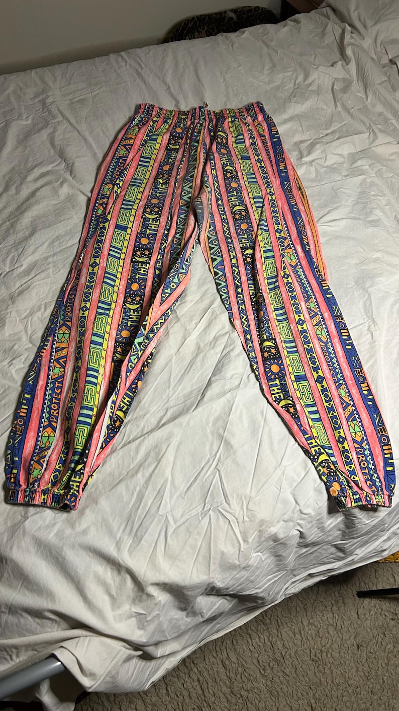 Puede incluir: Pantalones estampados con cintura con cord&oacute;n. El dise&ntilde;o presenta rayas verticales en rosa, amarillo, azul y verde, con dise&ntilde;os geom&eacute;tricos y solares. Los pu&ntilde;os el&aacute;sticos son visibles en los tobillos.