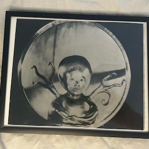 May include: A black and white framed print featuring a circular composition of a human-like figure with a large, rounded head and tentacle-like appendages extending from the shoulders, all enclosed within a dark, circular border.