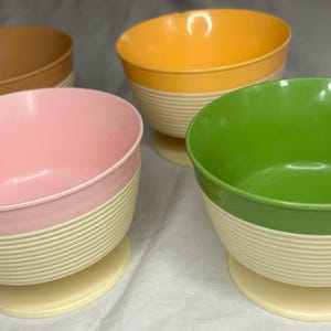 May include: Four colorful bowls in shades of brown, yellow, pink, and green. Each bowl has a ribbed, off-white base and a matching foot. The interiors of the bowls are the same color as the top rim.