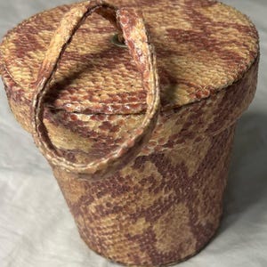 Puede incluir: Un bolso cilíndrico vintage con asa. El bolso está cubierto con un estampado de piel de serpiente en tonos beige, marrón y burdeos. El asa es de material de piel de serpiente a juego. El bolso tiene una parte superior circular y un pequeño cierre metálico.