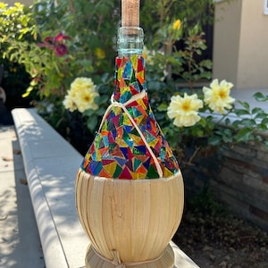 May include: A colorful mosaic glass bottle with a cork stopper and a woven straw covering. The bottle is sitting on a white surface.