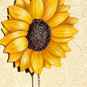 Könnte beinhalten: Ein gelber Sonnenblumen-Ausschnitt aus Holz mit brauner Mitte. Der Ausschnitt ist an einer silbernen Kette befestigt und dient als Hängedeko.