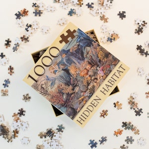 May include: A 1000 piece jigsaw puzzle with a colorful illustration of an underwater scene. The puzzle box is beige with the words "Hidden Habitat" printed on it.