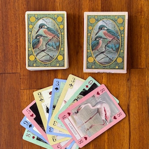 May include: Two decks of playing cards with a vintage bird illustration on the card backs. The face cards feature various illustrations, including a flamingo. The cards are arranged on a wooden surface.