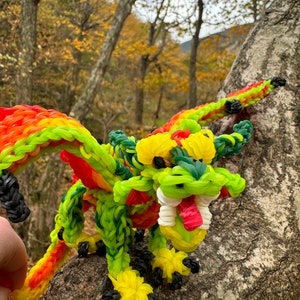 Peut inclure: Un dragon en élastique vert, jaune et rouge avec une langue blanche et rouge, perché sur un tronc d'arbre dans un environnement forestier.