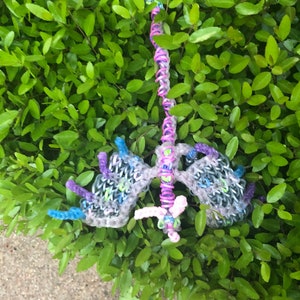 May include: A rainbow loom dragon made with colorful rubber bands. The dragon has a pink body, blue wings, and purple legs. It is sitting on a bed of green leaves.