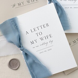 May include: A white card with the text "A Letter to My Wife on our wedding day July 30, 2022" and a blue ribbon tied around it.