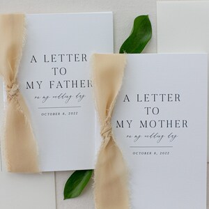 May include: Two white cards with black text, tied with beige ribbon. The text on the cards reads "A Letter to My Father on my wedding day October 8, 2022" and "A Letter to My Mother on my wedding day October 8, 2022".
