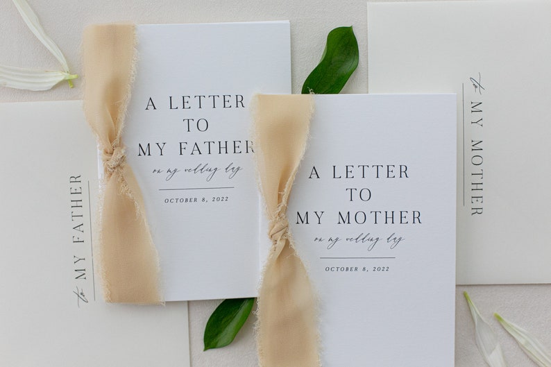 May include: Two white cards with black text, tied with beige ribbon. The text on the cards reads "A Letter to My Father on my wedding day October 8, 2022" and "A Letter to My Mother on my wedding day October 8, 2022".