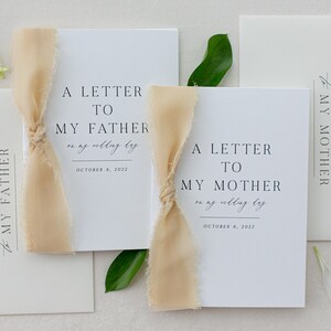 May include: Two white cards with a beige ribbon tied around them. The cards have the text "A Letter To My Father on my wedding day October 8, 2022" and "A Letter To My Mother on my wedding day October 8, 2022".