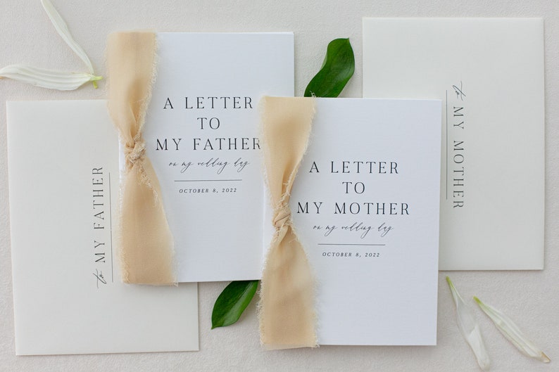 May include: Two white cards with a beige ribbon tied around them. The cards have the text "A Letter To My Father on my wedding day October 8, 2022" and "A Letter To My Mother on my wedding day October 8, 2022".