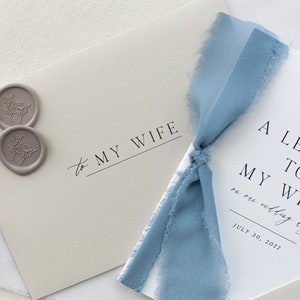 May include: A white card with the text "A Letter to My Wife on our wedding day July 30, 2022" and a blue ribbon tied around it. Two gray wax seals with floral designs are on the card to the left.