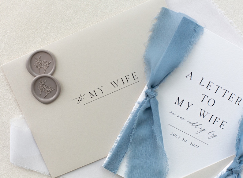 May include: A white card with the text "A Letter to My Wife on our wedding day July 30, 2022" and a blue ribbon tied around it. Two gray wax seals with floral designs are on the card to the left.