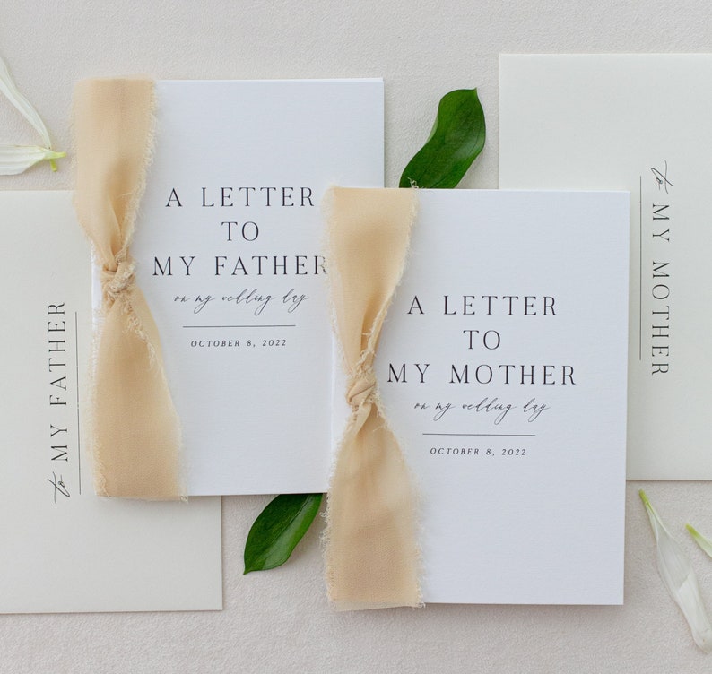 May include: Two white cards with a beige ribbon tied around them. The cards have the text "A Letter To My Father on my wedding day October 8, 2022" and "A Letter To My Mother on my wedding day October 8, 2022".