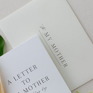 May include: A white card with the text "A Letter to My Mother" printed in black. The card is sitting on top of a cream-colored envelope with the text "to My Mother" printed in black.