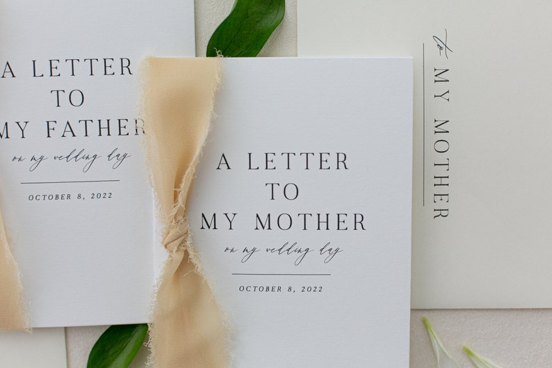 May include: Two white cards with the text "A Letter to My Father on my wedding day October 8, 2022" and "A Letter to My Mother on my wedding day October 8, 2022". A beige ribbon is tied around the cards.