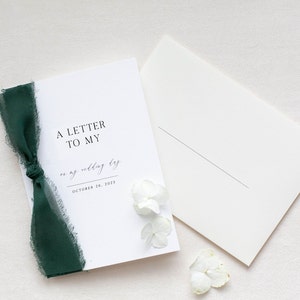 May include: A white card with the text "A Letter to My" and "on my wedding day" in cursive script. The date "October 28, 2023" is printed below the cursive text. A green ribbon is tied around the top left corner of the card. White flowers are scattered around the card.
