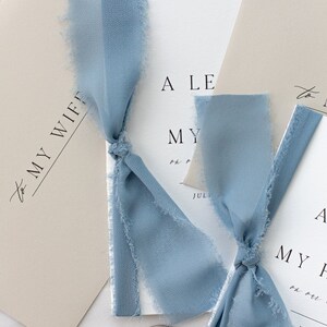May include: Two wedding invitations with a light blue ribbon tied around them. The invitations are on a white background with a light brown envelope and two wax seals.