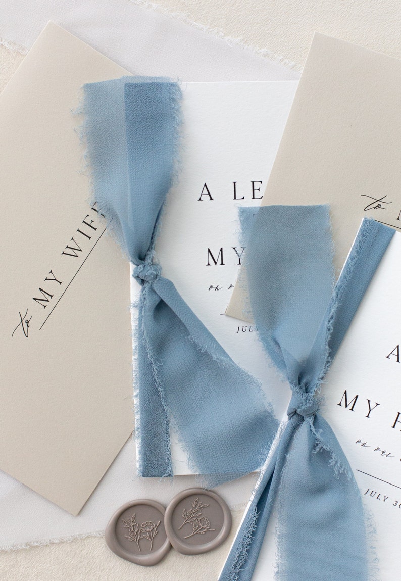 May include: Two wedding invitations with a light blue ribbon tied around them. The invitations are on a white background with a light brown envelope and two wax seals.