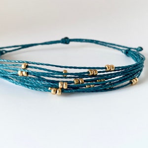 May include: A teal-colored bracelet featuring multiple strands of cord, each adorned with small, gold-colored beads. The bracelet is knotted at both ends, creating a simple yet elegant design. The jewelry is displayed on a white surface.