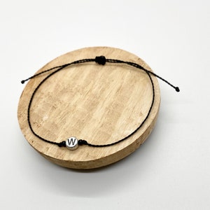 May include: A black string bracelet with a silver charm featuring the letter 'W'.