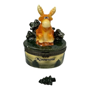May include: A ceramic trinket box featuring a sculpted orange moose figurine on the lid, surrounded by dark green trees. The base is cream-colored with a painted forest scene and the word Minnesota in black text. A small detached tree piece sits below.