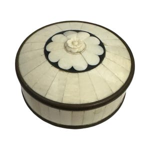 May include: A round lidded box featuring a radial pattern of off-white segments inlaid across the top and sides. The center of the lid displays a dark circular inlay with a carved white flower motif. The structure is framed by thin dark metal rims.