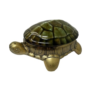 May include: A decorative turtle-shaped trinket box featuring a metallic gold-toned base with four legs and a head. The removable lid is crafted from glossy ceramic in shades of dark green and brown, displaying a textured, segmented shell pattern.