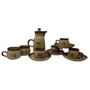 May include: A ceramic tea set featuring a tall lidded pitcher, a creamer, and four cups with matching saucers. Each piece has a cream-colored base with brown rim accents and a floral pattern. The text Denby is printed on the side of each cup.