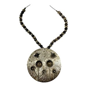 May include: Silver-toned necklace featuring a large, round pendant with a floral design. The pendant has a textured surface with embossed flowers and small butterfly accents. The necklace has a beaded chain.