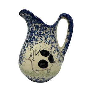 May include: A ceramic pitcher with a blue and white speckled design. The pitcher features a whimsical pig illustration with black spots. The handle is curved, and the spout is angled for easy pouring. A charming addition to any kitchen.