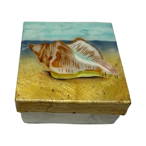 May include: A square decorative box with a seashell design. The box features a painted scene of a beach with a large seashell. The box has a gold-colored border and a white base.