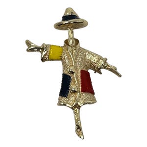 May include: A gold-tone metal brooch shaped like a scarecrow figure wearing a hat. The figure features a textured coat with a bright yellow sleeve, a dark blue panel, and a red panel, accented with small gold-tone buttons down the center.