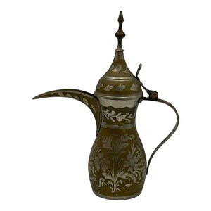 May include: A brass coffee pot with a long, curved spout, a hinged lid, and a tall, pointed finial. The body and lid feature intricate, etched silver-toned floral and leaf patterns that contrast against the muted, dark brass surface.