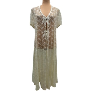 May include: A floor-length, cream-colored lace duster featuring short sleeves, a deep V-neckline, and a front closure with decorative floral appliqué details. The sheer fabric is adorned with an intricate, repeating embroidered vine pattern throughout.