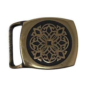 May include: A rectangular brass belt buckle featuring a rounded left side and a central circular inlay. The inlay displays a raised, intricate, symmetrical floral-inspired pattern in polished brass set against a dark, textured background.